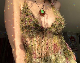 Puede incluir: Un top de crochet verde y marrón con flecos. El top se lleva sobre un top blanco sin mangas. La persona que lleva el top también lleva un collar con un colgante de piedra verde.