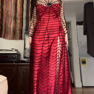 Könnte beinhalten: Ein langes, rotes Kleid mit einem schwarzen Spinnennetz-Design. Das Kleid hat einen hohen Ausschnitt und lange Ärmel mit einem Spinnennetz-Muster. Das Kleid hat einen Schlitz an der Seite. Das Kleid ist für Halloween oder Themenveranstaltungen geeignet.