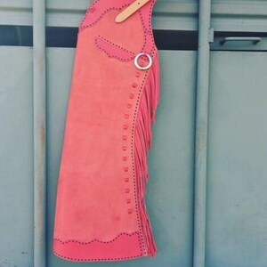 May include: A pink leather chaps with fringe and a brown leather strap. The chaps are hanging on a metal hook.