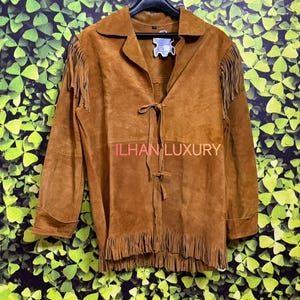 Könnte beinhalten: Braune Wildlederjacke mit Fransen an den Ärmeln und am Saum. Die Jacke hat einen Kragen und einen Bindverschluss in der Taille. Der Text "ILHAN LUXURY" ist auf der Vorderseite zu sehen.