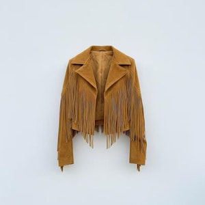 May include: A tan suede jacket featuring long fringe detailing across the chest, back, and sleeves. The jacket has a wide notched lapel collar, long sleeves, and a cropped hemline. The interior is lined with a matching tan fabric.