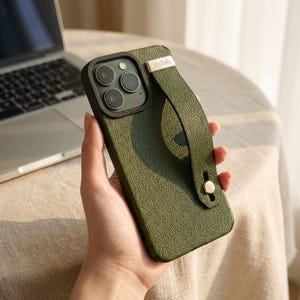 May include: Olive green phone case with a textured finish and a matching hand strap. The case has a black camera border and a small gold-colored metal accent that reads "stylish".