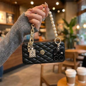 May include: Black quilted phone case with gold chain and pearl strap. The case features a gold rose emblem and a charm with the letter "a". The phone case has a camera cutout and is held by a person.