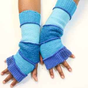 May include: A pair of blue arm warmers. The arm warmers are made of knitted fabric in shades of light blue, medium blue, and dark blue. The arm warmers have a ribbed cuff at the top and bottom. The arm warmers are designed to cover the forearm and leave the fingers exposed.