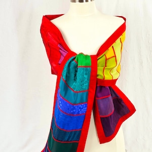 May include: A colorful patchwork scarf with a red border. The scarf is made of different fabrics in shades of red, orange, yellow, green, blue, purple, and black. The scarf is tied around a mannequin's neck.