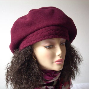 Puede incluir: Un gorro de lana de color burdeos con forro acolchado. El gorro se lleva en un maniquí con el pelo rizado marrón.