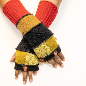 May include: A pair of fingerless gloves with a patchwork design. The gloves feature red ribbed cuffs, with sections of mustard yellow, black, and textured gold fabric. The edges are trimmed with black and red stitching.