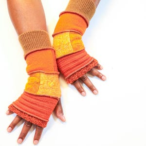May include: A pair of handmade arm warmers in shades of orange and yellow. The arm warmers feature a patchwork design with various textures, including knitted and felted sections. The cuffs are a darker brown color.