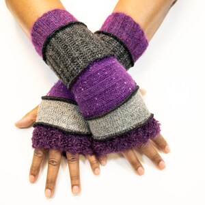 May include: A pair of knitted fingerless gloves in shades of purple, gray, and black. The gloves have a textured design with a ribbed cuff and a fluffy trim. The gloves are displayed on a white background.