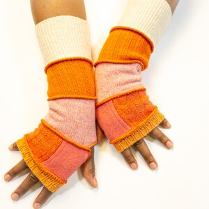 May include: A pair of colorful fingerless gloves. The gloves feature a cream-colored cuff, followed by sections of orange and pink knit fabric. The design includes cable knit and ribbed textures. The gloves are displayed against a white background.