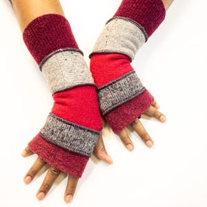 May include: A pair of handmade fingerless gloves in shades of red, gray, and burgundy. The gloves feature a layered design with ribbed cuffs and a textured appearance. The gloves are designed to cover the hands and wrists, leaving the fingers exposed.