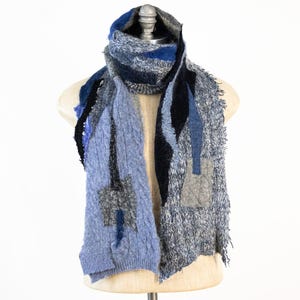 Może przedstawiać: Szalik w kolorze niebieskim, szarym i czarnym z frędzlami. Szalik jest wykonany z miękkiej, puszystej przędzy i ma patchworkowy wzór.