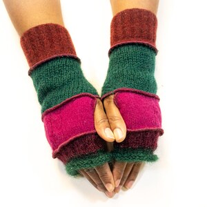 May include: A pair of handmade fingerless gloves in shades of green, burgundy, and fuchsia. The gloves are knitted with a ribbed cuff and a contrasting band of color at the wrist and mid-hand. The gloves are displayed against a white background.