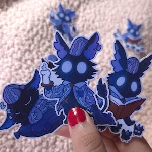 May include: A collection of blue and purple stickers featuring cartoon creatures with large eyes and patterned clothing. One holds a candle, another a book. The stickers are held in a hand with red nail polish.