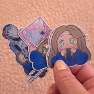 May include: A collection of colorful stickers featuring cartoon illustrations. One sticker depicts a girl with large eyes holding a phone. Other stickers show a futuristic soldier and a drawing of a blue cat. The stickers are held by a hand.