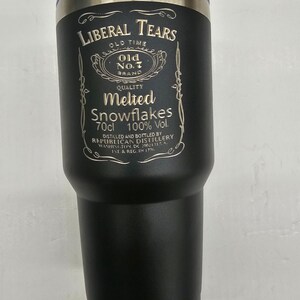 May include: A black stainless steel tumbler with a gold-colored design that reads "Liberal Tears Old Time Old No.7 Brand Quality Melted Snowflakes 70cl 100% Vol. Distilled and Bottled by Republican Distillery Washington, DC 20024 USA. 1st & Reg. In 1776."