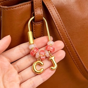 May include: A gold-toned U-shaped carabiner keyring featuring a series of translucent pink and white beads, accented with two polished gold-toned metal letter charms, C and J, hanging from the bottom of the bead arrangement.