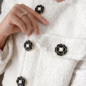 May include: Close-up of a white tweed jacket featuring black flower-shaped buttons. The buttons have a gold trim and a white centre. The jacket has a textured appearance and a classic design.