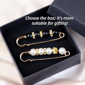 Könnte beinhalten: Zwei dekorative Sicherheitsnadeln mit goldfarbenem Metall und Perlenakzenten, eine mit schwarzen Perlen und die andere mit weißen Perlen, werden in einer schwarzen Geschenkbox präsentiert. Der Text lautet: Wähle die Box; sie eignet sich besser zum Verschenken!