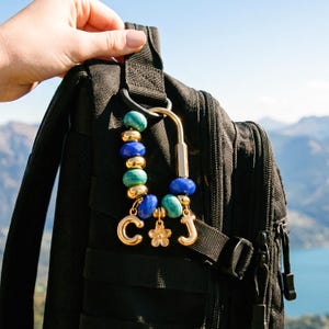 Può includere: Un portachiavi a moschettone color oro con una serie di perline turchesi e blu reale alternate a sfere color oro. Tre ciondoli color oro pendono dalla parte inferiore, tra cui le lettere C e J e una piccola forma di fiore.