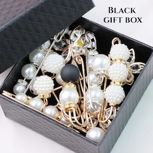 May include: A black gift box filled with decorative pins. The pins feature pearls, crystals, and gold-colored metal accents. Some pins have a black spherical bead. The box is open, revealing the contents.