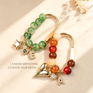 Puede incluir: Dos llaveros de mosquetón de metal dorado con cuentas. Uno tiene cuentas verdes, un dije de estrella dorada, un corazón verde y la letra E. El otro tiene cuentas naranjas y crema, un dije de corazón dorado, un corazón pequeño y la letra A. Texto: 1. CHOOSE BIRTHSTONE 2. CHOOSE YOUR INITIAL