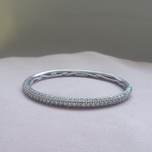Op de afbeelding: Een zilverkleurige scharnierende armband met een pavé-gezette buitenkant versierd met talloze kleine, heldere, sprankelende steentjes. De binnenband toont een zichtbaar keurmerk en het ontwerp bevat een veilig sluitingsmechanisme.