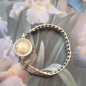 May include: A silver-toned wristwatch with a round, champagne-colored dial featuring the Omega logo and the word Ladymatic. The watch has a flexible, expandable metal link bracelet with a textured pattern on the links near the watch case.