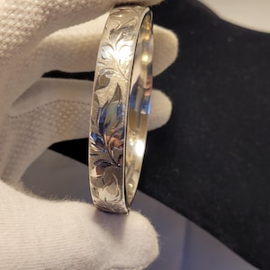 Può includere: Un bracciale rigido in metallo color argento caratterizzato da una superficie testurizzata con un motivo a foglie inciso. L'esterno della fascia presenta un disegno botanico ripetuto con riflessi lucidi su uno sfondo opaco.