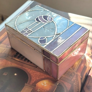 May include: A rectangular stained glass box with a light blue, purple, and pink design. The box has a gold-colored metal trim and is sitting on a book with a vintage architectural image.