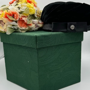 May include: A green gift box with a black velvet hat and a floral hat on top. The floral hat has orange, yellow, and white flowers.