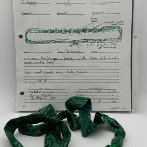 May include: A green paisley fabric necklace with green glass beads. The necklace is 14 inches long and the beads are 1 inch wide. The necklace is labeled "GG.97.04.06.PBK" and "Necklace, paisley cloth tube alternately bead marble bead".