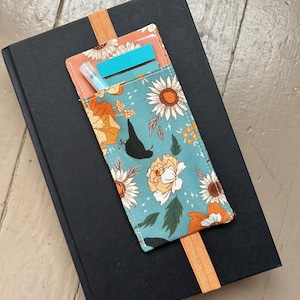 May include: A black book with a teal fabric pen holder featuring a floral pattern of sunflowers and other flowers in orange and white. The pen holder has a clear pocket with a blue sticky note and a pen. An orange elastic band holds the pen holder to the book.
