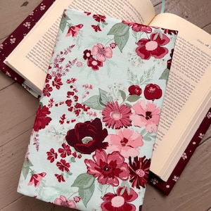 May include: A floral book cover with a light blue background and a pattern of red and pink flowers. The book cover is open, revealing the pages of a book. The cover is made of fabric.