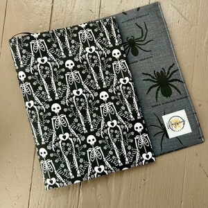 Puede incluir: Una funda de tela para libro con fondo negro y estampado de esqueletos y flores blancas, sobrepuesta a una funda gris con estampado de ara&ntilde;as negras y una etiqueta cuadrada blanca con el texto Mayberry Arts impreso en negro y amarillo.