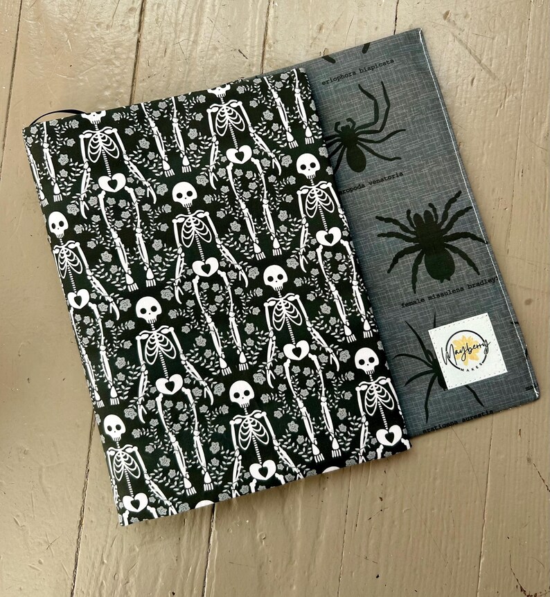 Puede incluir: Una funda de tela para libro con fondo negro y estampado de esqueletos y flores blancas, sobrepuesta a una funda gris con estampado de ara&ntilde;as negras y una etiqueta cuadrada blanca con el texto Mayberry Arts impreso en negro y amarillo.
