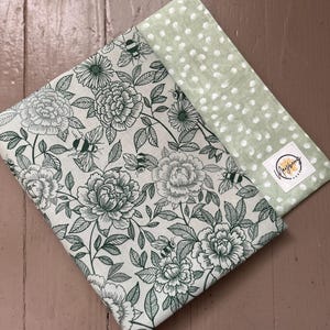 May include: A fabric book cover and bookmark set. The book cover features a floral and bee pattern in green and white. The bookmark is light green with white polka dots. The set is on a wooden surface.
