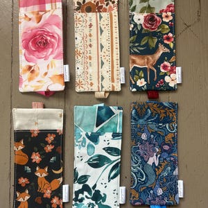 May include: Six rectangular fabric sleeves with elastic pull tabs, each featuring distinct patterns including pink roses, geometric shapes, a deer in a floral forest, foxes with flowers, teal watercolor leaves, and a mermaid with sea creatures.