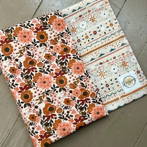 May include: Two fabric pieces with floral and patterned designs. One features large peach and brown flowers on a white background. The other has a cream background with geometric and floral patterns in peach, brown, and teal. A small tag with the word "Mayberry" is visible.