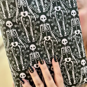Puede incluir: Un art&iacute;culo de tela rectangular con un patr&oacute;n repetitivo de esqueletos blancos y peque&ntilde;as rosas grises sobre un fondo negro. Los esqueletos se muestran en posici&oacute;n de pie con huesos p&eacute;lvicos en forma de coraz&oacute;n, dispuestos verticalmente.