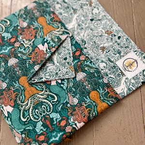 May include: A green fabric envelope with a colorful sea creature pattern. The envelope features mermaids, octopuses, and seashells. The envelope is folded in half and has a white label with the text "Mayberry" on it.