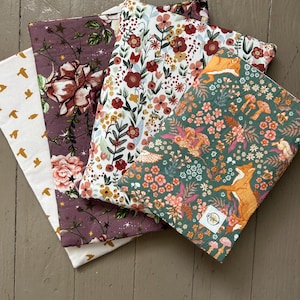 May include: A stack of four fabric items with floral and nature-themed patterns. The fabrics feature designs with flowers, birds, and foxes in various colors, including white, purple, and green. The items are arranged on a wooden surface.