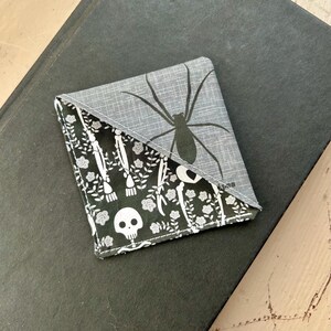 Puede incluir: Un marcap&aacute;ginas de esquina de tela triangular con una secci&oacute;n superior gris con un gr&aacute;fico de ara&ntilde;a negra y una secci&oacute;n inferior negra con un patr&oacute;n blanco de calaveras, huesos y peque&ntilde;as flores. La tela tiene costuras visibles en los bordes.