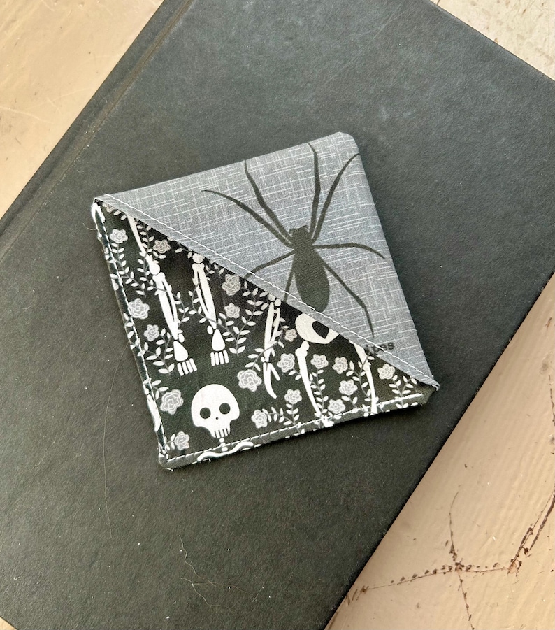 Puede incluir: Un marcap&aacute;ginas de esquina de tela triangular con una secci&oacute;n superior gris con un gr&aacute;fico de ara&ntilde;a negra y una secci&oacute;n inferior negra con un patr&oacute;n blanco de calaveras, huesos y peque&ntilde;as flores. La tela tiene costuras visibles en los bordes.