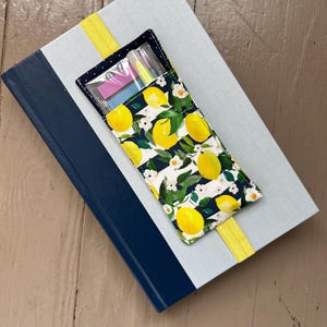 May include: A light blue book with a navy blue spine and a yellow ribbon bookmark. A pencil case with a lemon pattern rests on top, containing highlighters and sticky notes. The pencil case is navy blue with a lemon and floral print.