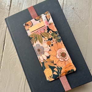 May include: A floral print pen holder with a dark background, resting on a dark book. The pen holder features a colorful floral pattern with orange, pink, and green hues. A pink ribbon bookmark is attached to the book.