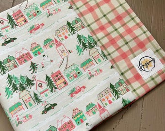 May include: A fabric book cover featuring a whimsical Christmas village pattern with houses, trees, and cars in red, green, and pink on a cream background. The interior lining is a pink and green plaid. A small label with the text "Mayberry Makes" is visible.