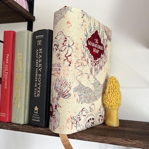 May include: A white book cover with a red and black map design. The map is titled "The Marauder's Map". The book is on a wooden shelf with other books.