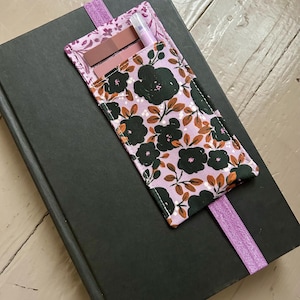 May include: A floral print bookmark with a pocket, holding a pen and two small rectangular items, rests on a closed black book. The bookmark features a lavender background with dark green flowers and brown leaves. A lavender elastic band is attached.