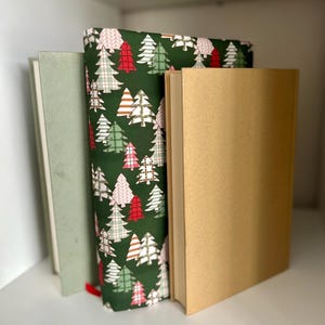 May include: Three books standing upright. The center book features a dark green fabric cover with a repeating pattern of stylized Christmas trees in shades of red, pink, white, and light green. Flanking it are a light sage green book and a tan book.